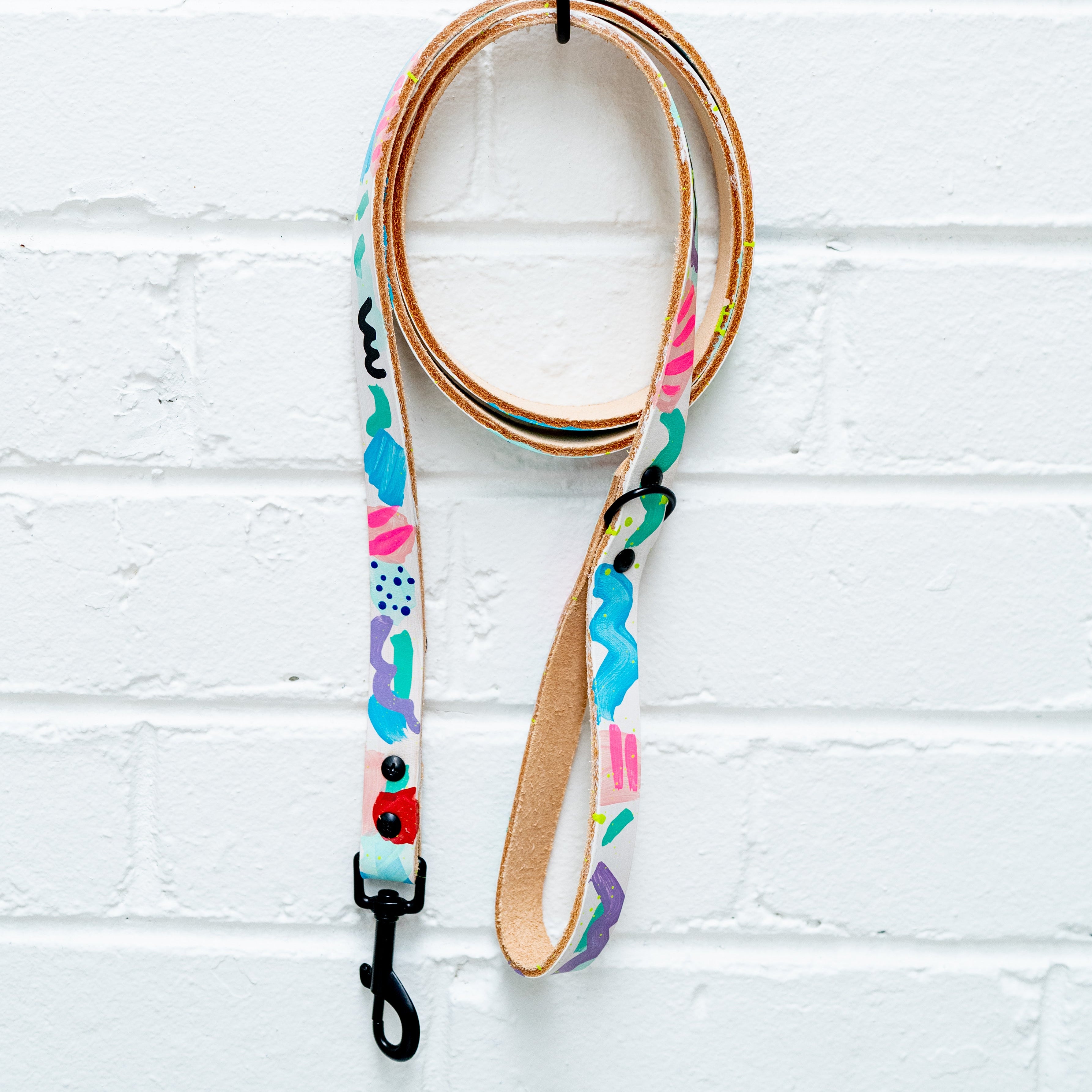 Colorful dog leash with patterns hanging on a palm tree hook against a white brick wall.