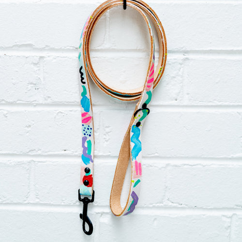 Colorful dog leash with patterns hanging on a palm tree hook against a white brick wall.